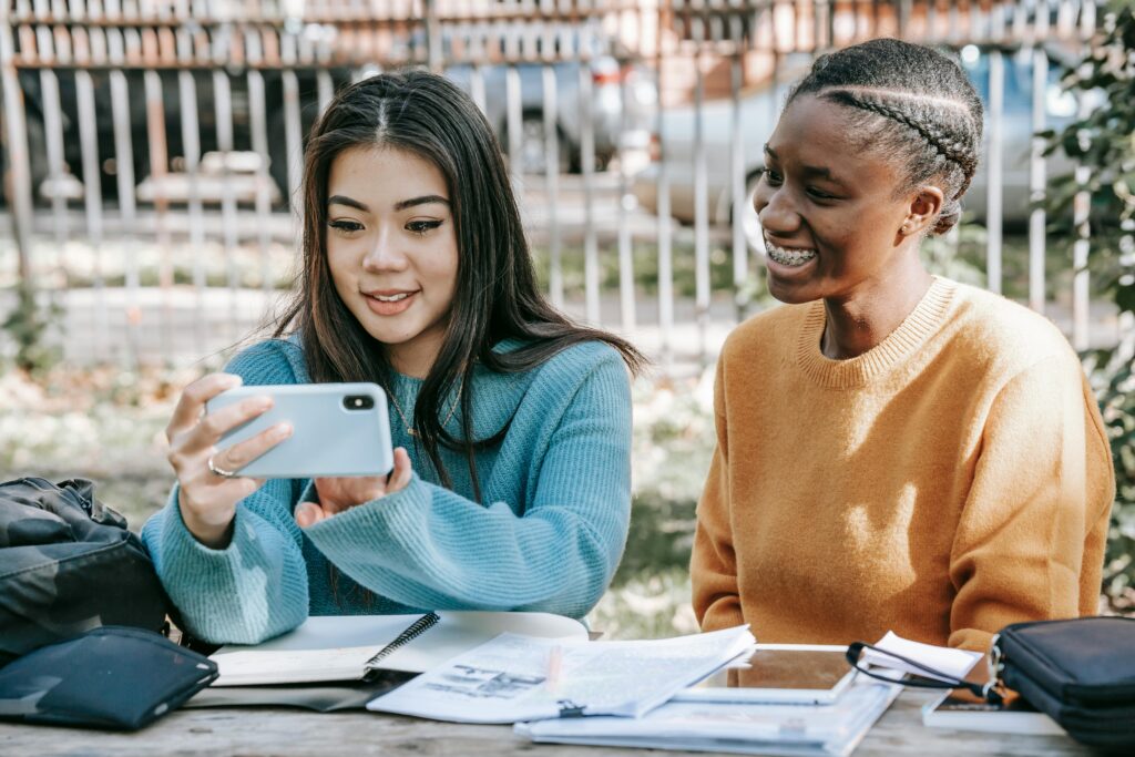 Two smiling female students sitting outdoors, looking at a smartphone together, with notebooks and study materials on the table, reflecting the impact of social media on student life.